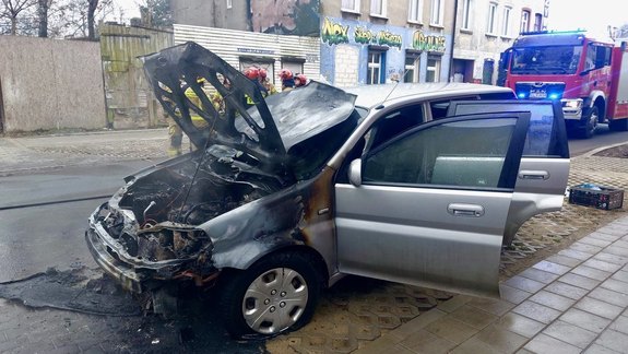 Spalone auto, w tle strażacy w trakcie akcji i wóz gaśniczy.