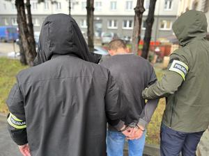 Policjanci trzymają mężczyznę, który ma kajdanki założone na ręce z tyłu.