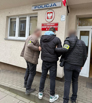 Policjanci prowadzą mężczyznę w kajdankach zespolonych.