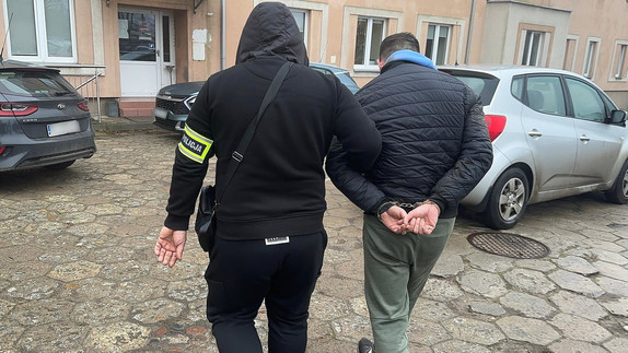 Policjant prowadzi mężczyznę, który ma założone kajdanki na ręce z tyłu.