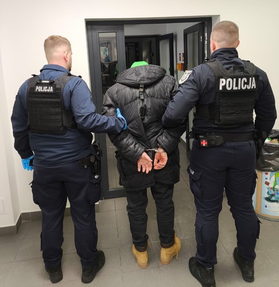 Policjanci trzymają mężczyznę, który ma kajdanki założone na ręce z tyłu.
