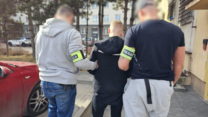 Policjanci trzymają mężczyznę, który ma założone kajdanki zespolone.