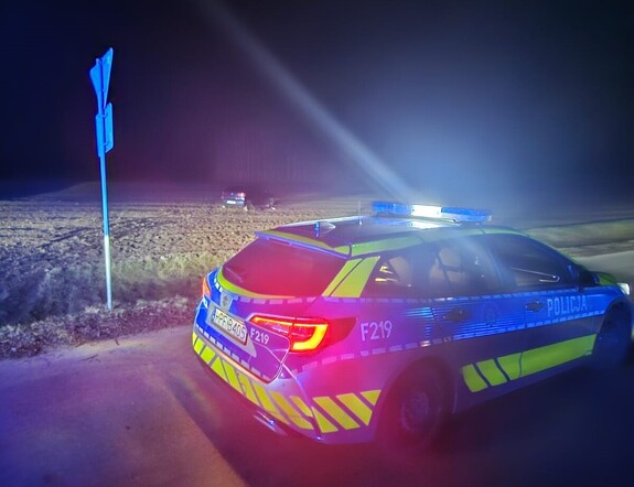 Oznakowany radiowóz policyjny z włączonymi sygnałami świetlnymi, w  tle - poza drogą - stoi pojazd.