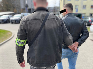 Policjant prowadzi mężczyznę, który ma założone kajdanki na ręce z tyłu.