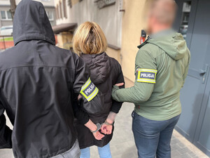 Policjanci trzymają kobietę, która ma założone kajdanki na ręce z tyłu.