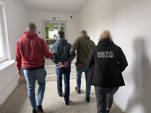 Policjanci prowadzą mężczyznę, który ma założone kajdanki na ręce z tyłu.