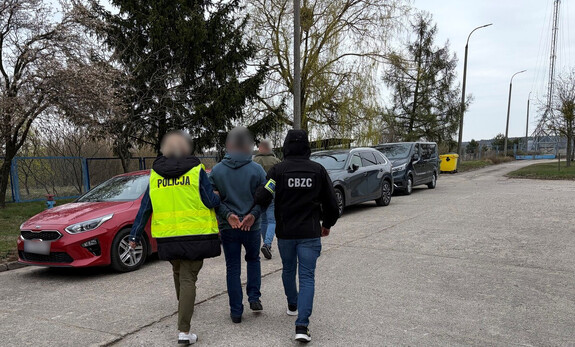 Policjanci prowadzą mężczyznę, który ma założone kajdanki na ręce z tyłu.