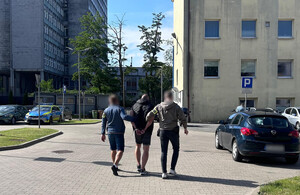 Policjanci prowadzą mężczyznę, który ma założone kajdanki na ręce z tyłu.