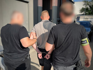 Policjanci prowadzą mężczyznę, który ma założone kajdanki na ręce z tyłu.