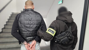 Policjant trzyma mężczyznę, który ma założone kajdanki na ręce z tyłu.