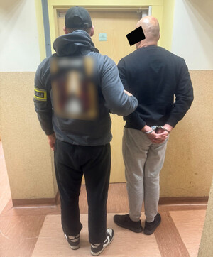 Policjant trzyma mężczyznę, który ma założone kajdanki na ręce z tyłu.