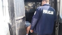 Na zdjęciu policjant, który stoi przy otwartych drzwiach radiowozu (tylnych). W środku we wnętrzu pojazdu sierdzi mężczyzna w kajdankach.
