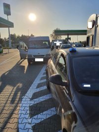 Na zdjęciu dwa pojazdy stojące na poboczu w pobliżu stacji benzynowej. Policjant ruchu drogowego stoi przy kontrolowanym pojeździe typu bus. Widoczny jest też na zdjęciu nie oznakowany radiowóz Policji.