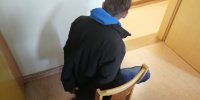 Na zdjęciu widoczny jest mężczyzna siedzący na krześle bokiem do obiektywu aparatu. Mężczyzna ma założone na rękach kajdanki. Znajduje się w pomieszczeniu. Ubrany jest w ciemną kurtkę spod której widać wystający niebieski kaptur.