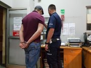 Na zdjęciu widać mężczyznę z zapiętymi na dłoniach kajdankami oraz policjanta w granatowym mundurze