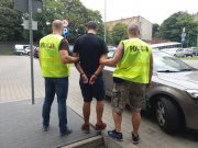 Na zdjęciu widzimy wysokiego mężczy8znę , ubranego w czarną koszulkę i spodenki do kolan, ma z tyłu zakute ręce w kajdanki, po jego obu stronach stoi dwóch policjantów po cywilnemu w zielonych kamizelkach policyjnych.