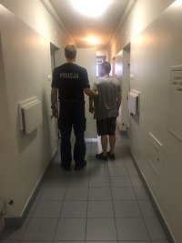 Zdjęcie jest zrobione na Komisariacie w Zgierzu. Widać na nim stojącego mężczyznę na korytarzu , tyłem odwróconego do aparatu. Trzyma go policjant, który jest ubrany w mundur policyjny.