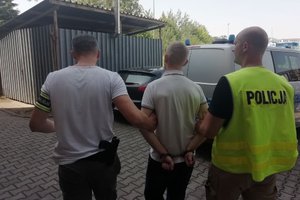 Dwóch policjantów prowadzi mężczyznę z kajdankami założonymi na ręce trzymane z tyłu