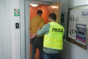 Policjant w kamizelce z napisem Policja wchodzi do windy z mężczyzną z kajdankami na rękach z tyłu