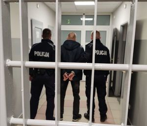 Dwóch policjantów i mężczyzna z kajdankami na rękach stoją za kratą.