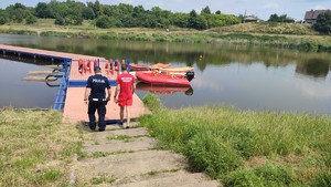 Na zdjęciu widać zalew Zadębie, pomost, na który wchodzą policjant i ratownik WOPR. Zmierzają do łódki.