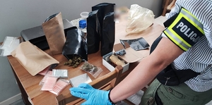 Na pierwszym planie ręka z opaską na wysokości łokcia z napisem POLICJA, dalej różnego rodzaju narkotyki ułożone na biurku.