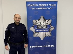 Umundurowany Policjant na tle logo Komendy Miejskiej Policji w Skierniewicach