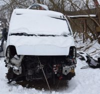 Na zdjęciu samochód typu bus koloru białego uszkodzony zasypany śniegiem. Pojazd nie posiada przednich elementów karoserii widać uszkodzony silnik.