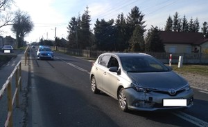 Na jezdni stoją dwa osobowe samochody, między nimi oznakowany radiowóz z włączonym światłami błyskowymi, za barierkami ochronnymi na poboczu zaparkowany samochód osobowy.