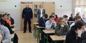 W klasie szkolnej stoi jeden umundurowanych policjant, jedna kobieta, przed nim w ławkach siedzą uczniowie.