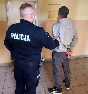 W pomieszczeniu, na korytarzu stoi tyłem umundurowany policjant, który trzyma mężczyzną stojącego również tyłem zapiętego w kajdanki na ręce trzymane w tyle.