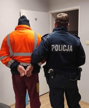 Policjant i mężczyzna z kajdankami na rękach trzymanymi w tyle odwróceni plecami.