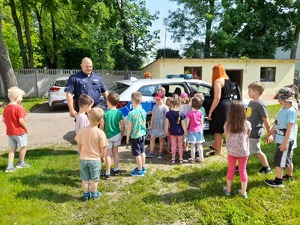 Grupa dzieci ogląda oznakowany radiowóz, po lewej stronie stoi uśmiechnięty policjant.