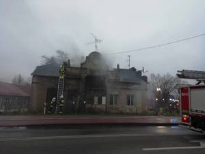 Budynek z którego wydobywa się dym i strażacy gaszący pożar.