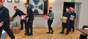Policjanci i pracownik cywilny Policji niosący pod choinkę prezenty. Wszyscy mają czerwone czapki z białym pomponem.