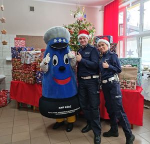 Policjant, policjantka i maskotka garnizonu łódzkiego podnoszą kciuk do góry i pozują do wspólnego zdjęcia. W tle stół na którym leżą paczki z przezentami.