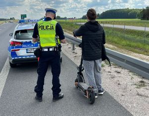 Policjant stoi obok kierującego hulajnogą. Obaj znajdują się na drodze ekspresowej. Za nimi widać oznakowany radiowóz.