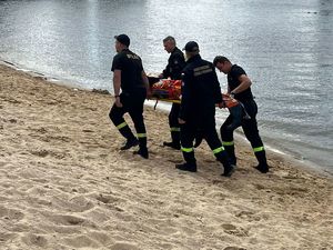 Strażacy na noszą niosą pokrzywdzonego. Funkcjonariusze idą przy piaszczystym brzegu.