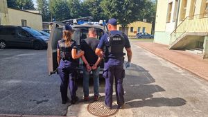 Policjanci stoją przed radiowozem z zatrzymanym mężczyzną.