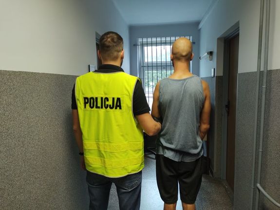 Policjant i mężczyzna stoją tyłem. Policjant trzyma mężczyznę za rękę.