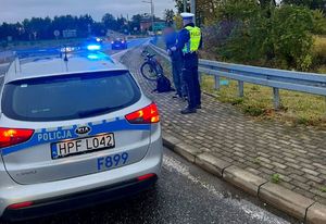 Policyjny radiowóz. W tle policjant wykonujący czynności służbowe z rowerzystą znajdującym się na drodze ekspresowej.