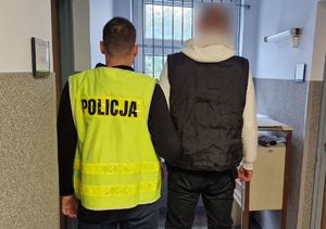 Policjant stoi z zatrzymanym mężczyzną. Funkcjonariusz jest nieumundurowany, posiada na sobie kamizelkę odblaskową z napisem Policja.