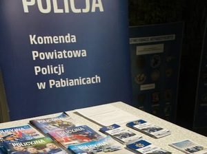 Stoisko doborowe na którym widać stolik z gazetami policyjnymi i ulotkami informacyjnymi. W tle stoi baner z napisem: Komenda Powiatowa Policji w Pabianicach.