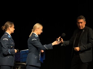 Na scenie stoją dwie policjantki, jedna z nich przekazuje mikrofon mężczyźnie.