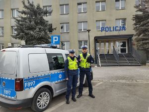 stojący policjant z policjantką przy radiowozie przed komendą Policji