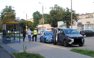 Miejsce zdarzenia drogowego znajdujące się w pobliżu przystanku autobusowego. Na zdjęciu widać osoby uczestniczące w zdarzeniu, policjanta, radiowóz oraz inne pojazdy.
