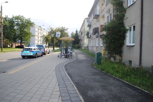 Miejsce zdarzenia drogowego znajdujące się w pobliżu przystanku autobusowego. Na zdjęciu widać osoby uczestniczące w zdarzeniu, radiowóz oraz inne pojazdy.