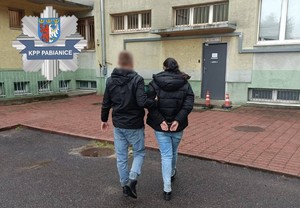 Nieumundurowany policjant idzie z zatrzymaną kobietą. W lewym górnym rogu znajduje się logo Komendy Powiatowej Policji w Pabianicach.