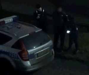 Policjanci stoją z zatrzymanym mężczyzną przy radiowozie.