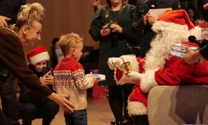 Chłopiec odbiera czekoladki od Świętego Mikołaja, za nim stoi pochylona kobieta.